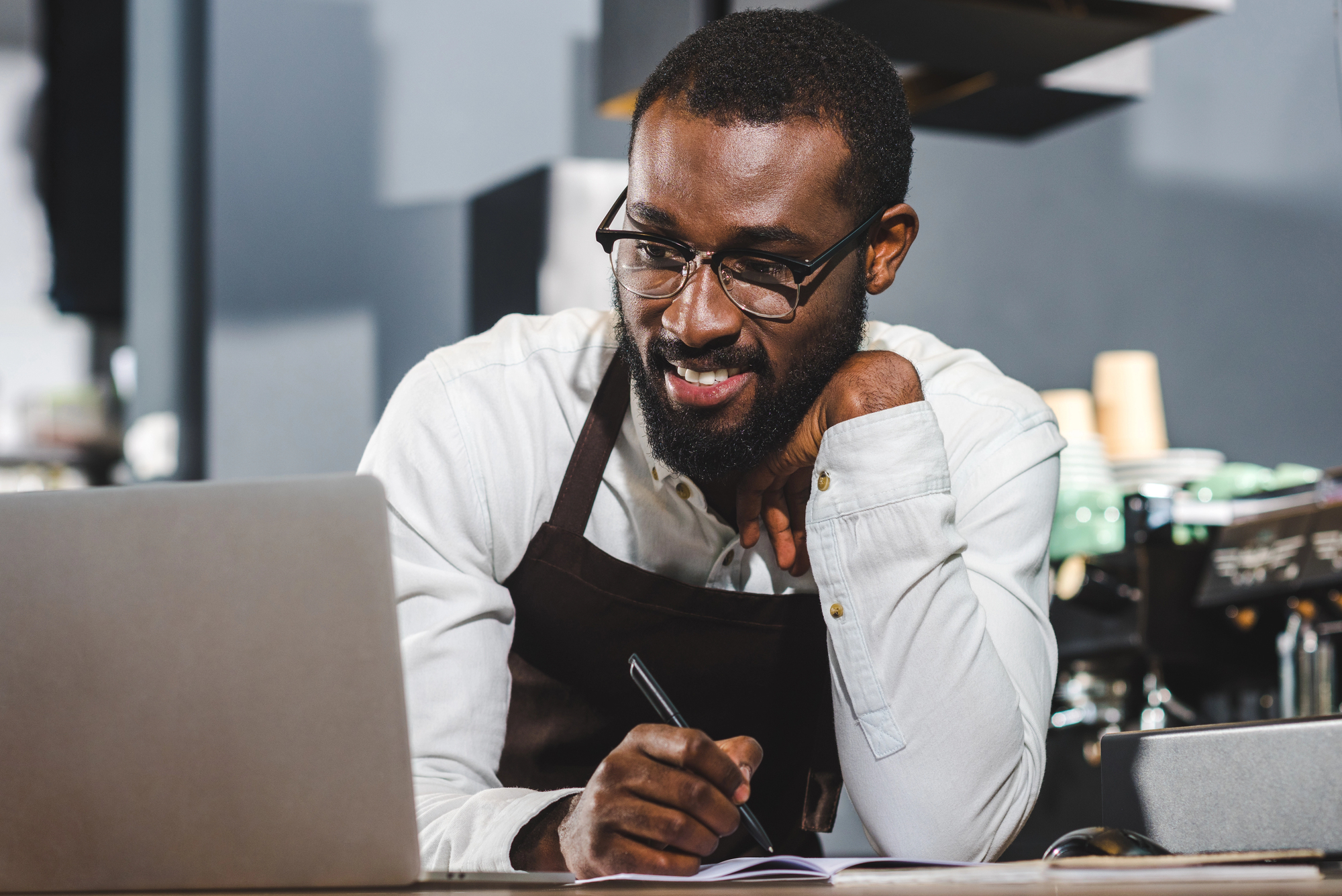 A man wearing glasses and an apron is smiling while looking at a laptop. He is holding a pen and writing in a notebook in front of him. There is a coffee machine in the background.