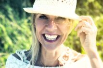 A woman with bright lipstick smiles warmly, holding a woven hat on her head. She's outdoors with green foliage in the background, wearing a light-colored top. Sunlight touches her face, creating a cheerful and vibrant atmosphere.