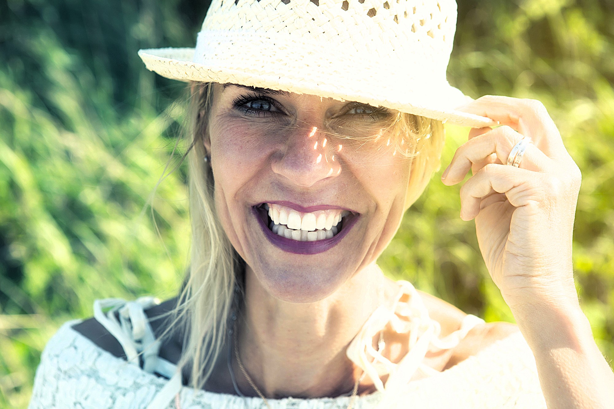 A woman with bright lipstick smiles warmly, holding a woven hat on her head. She's outdoors with green foliage in the background, wearing a light-colored top. Sunlight touches her face, creating a cheerful and vibrant atmosphere.