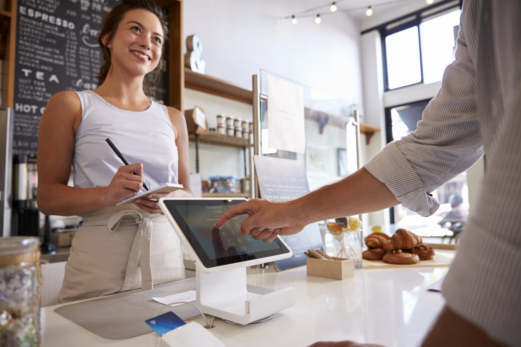 A person is using a touchscreen register at a cafe, placing an order. A smiling barista stands behind the counter, holding a notepad. Fresh pastries are displayed nearby, and the cafe interior is bright with natural light.