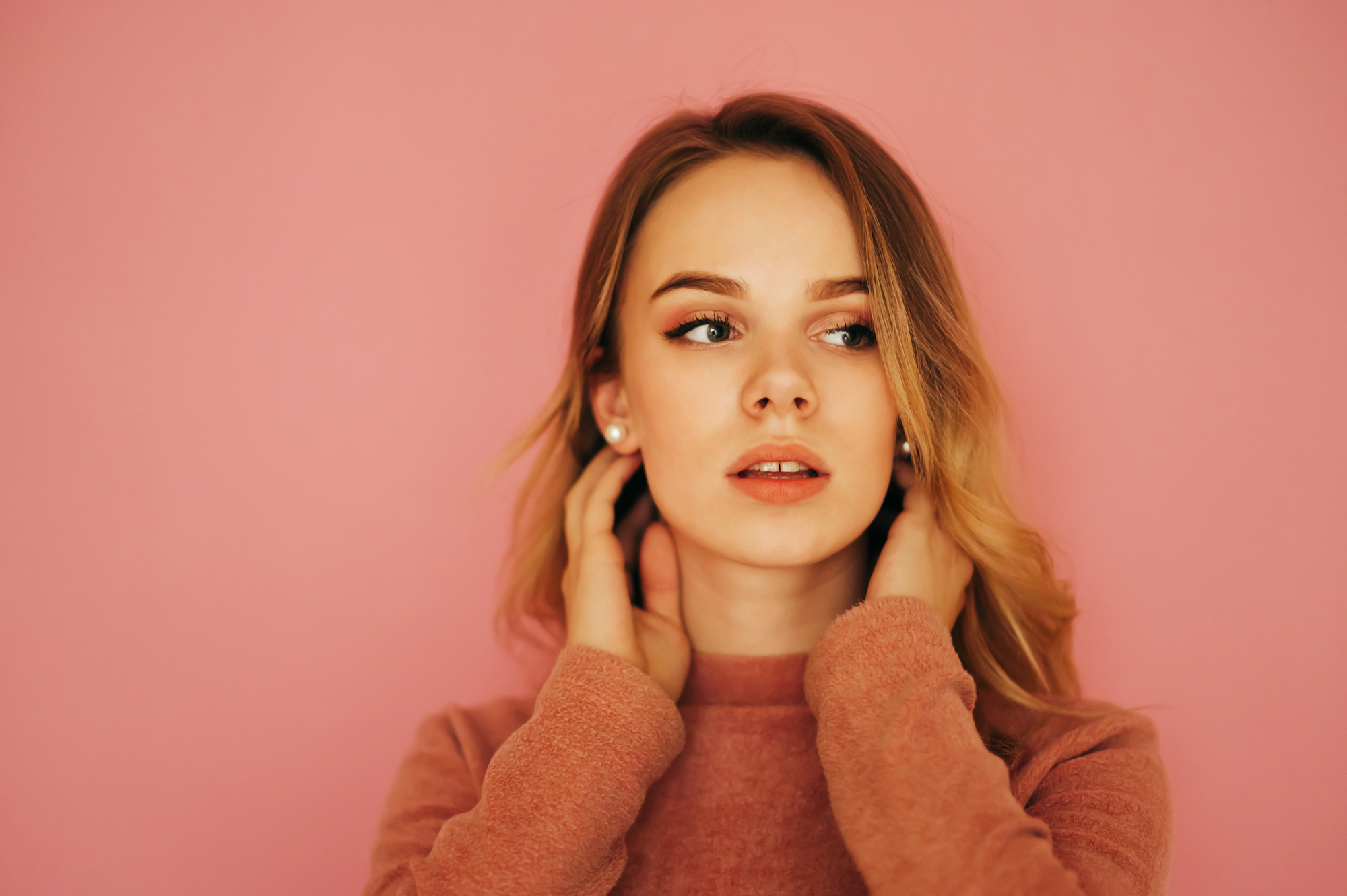 A young woman with long blonde hair poses against a pink background. She looks slightly to the side, with her hands near her face and wears a peach-colored sweater.