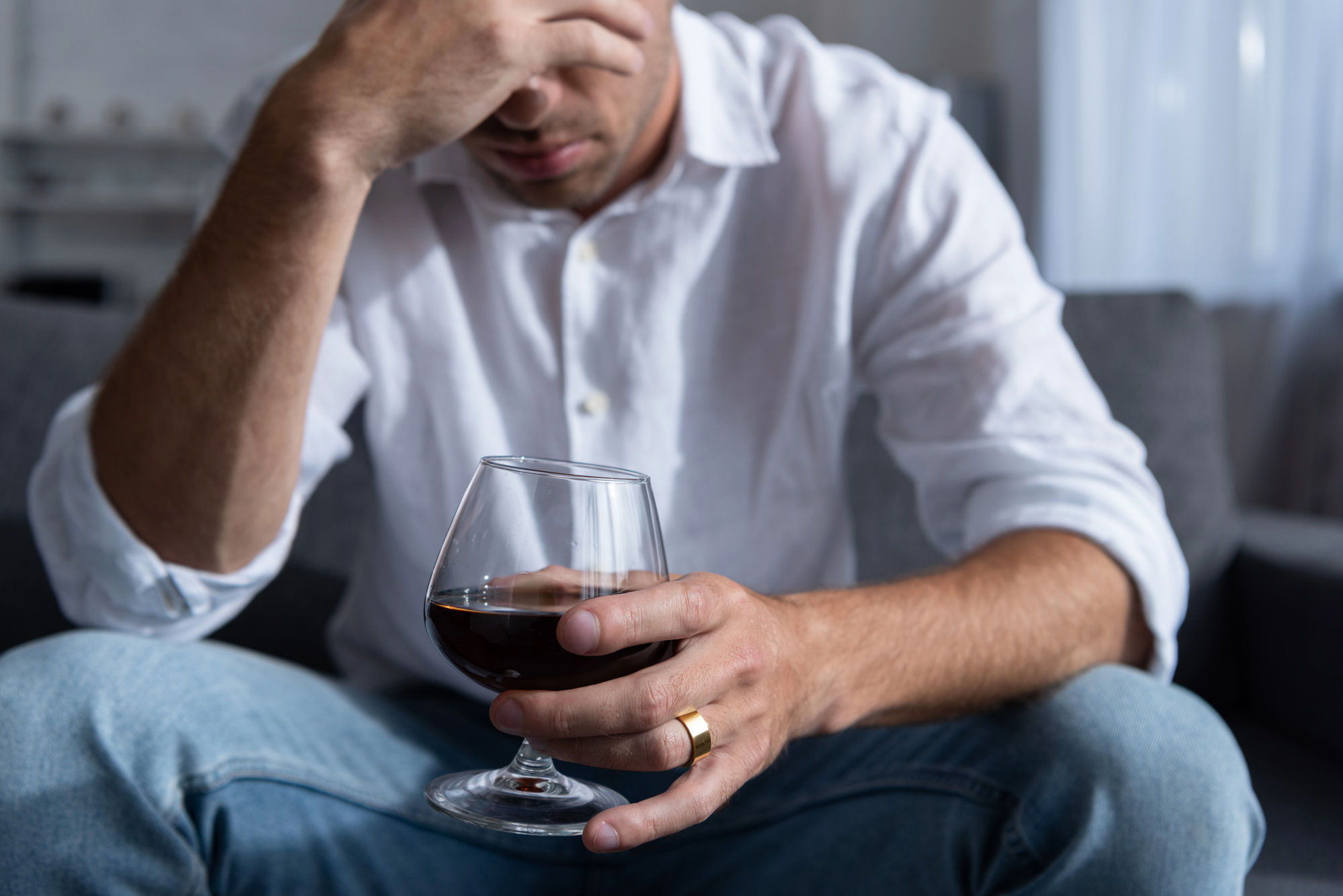 A man wearing a white shirt and jeans sits on a couch, holding a glass of dark liquid in one hand. His other hand rests on his forehead, and he wears a gold ring. The background is blurred, suggesting a home setting.