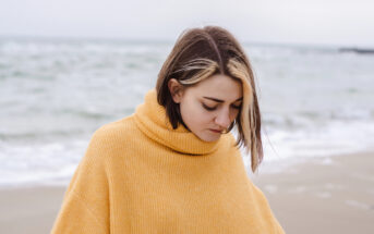 A person with shoulder-length hair and a blonde streak stands on a beach, wearing a mustard yellow turtleneck sweater. The sea is in the background under a cloudy sky. The person gazes downwards, creating a calm and introspective mood.