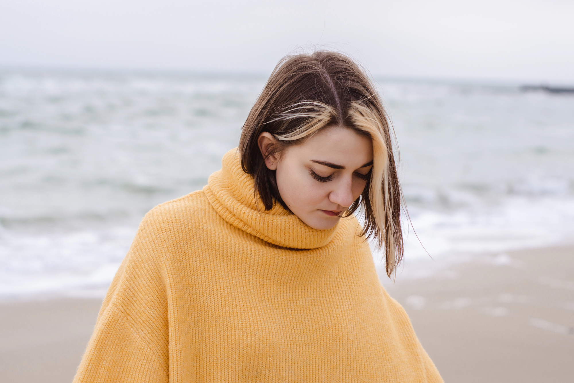 A person with shoulder-length hair and a blonde streak stands on a beach, wearing a mustard yellow turtleneck sweater. The sea is in the background under a cloudy sky. The person gazes downwards, creating a calm and introspective mood.