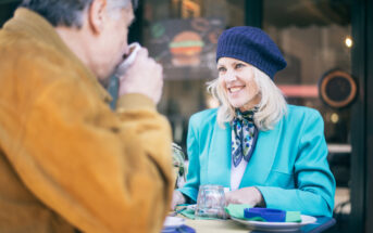 A woman in a teal blazer and navy beret smiles at a man in a brown jacket as they sit at an outdoor cafe table. The man sips from a cup. Green table settings are visible. The background features a blurred cafe sign.