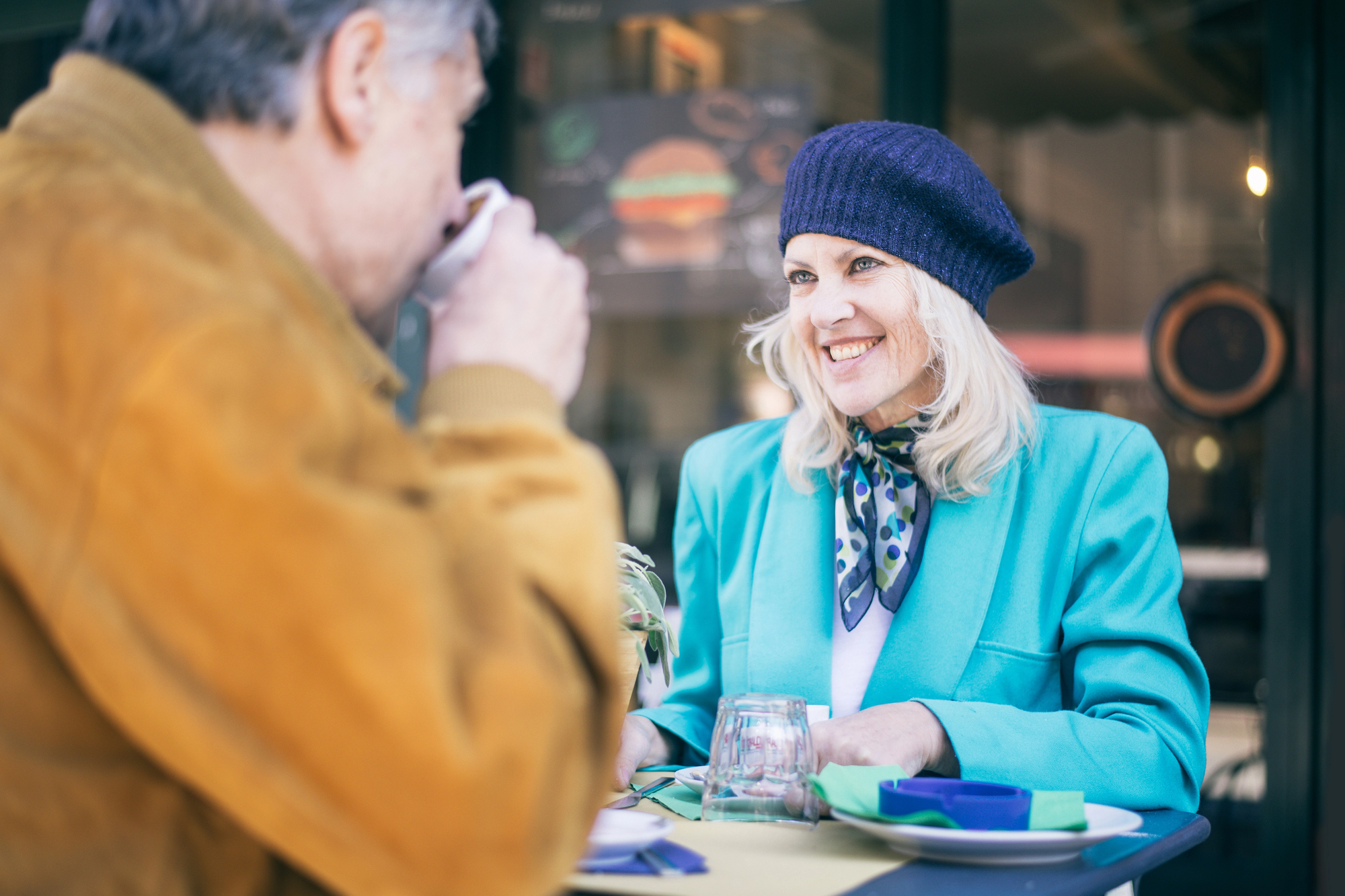 A woman in a teal blazer and navy beret smiles at a man in a brown jacket as they sit at an outdoor cafe table. The man sips from a cup. Green table settings are visible. The background features a blurred cafe sign.