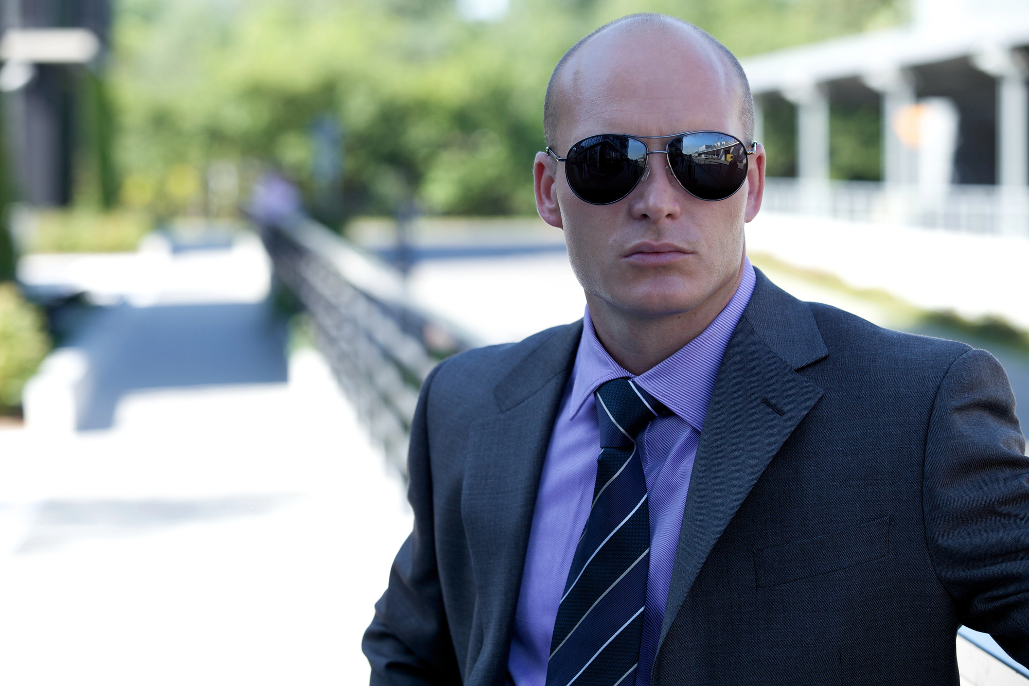 A bald man in a grey suit and purple shirt wearing sunglasses stands outside in a sunlit area. The background features trees and a blurry building walkway.