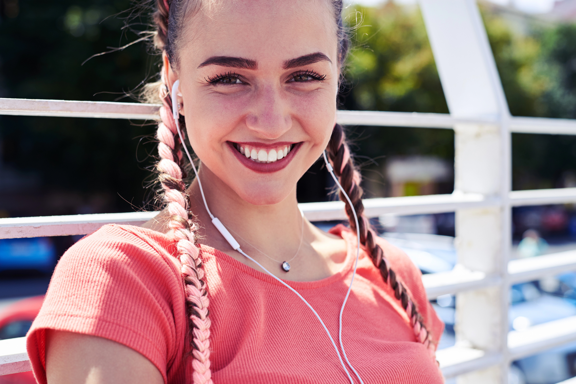 A woman with braided hair and wearing earphones smiles brightly at the camera. She is wearing a peach-colored top and standing against a white railing with blurred greenery and cars in the background.