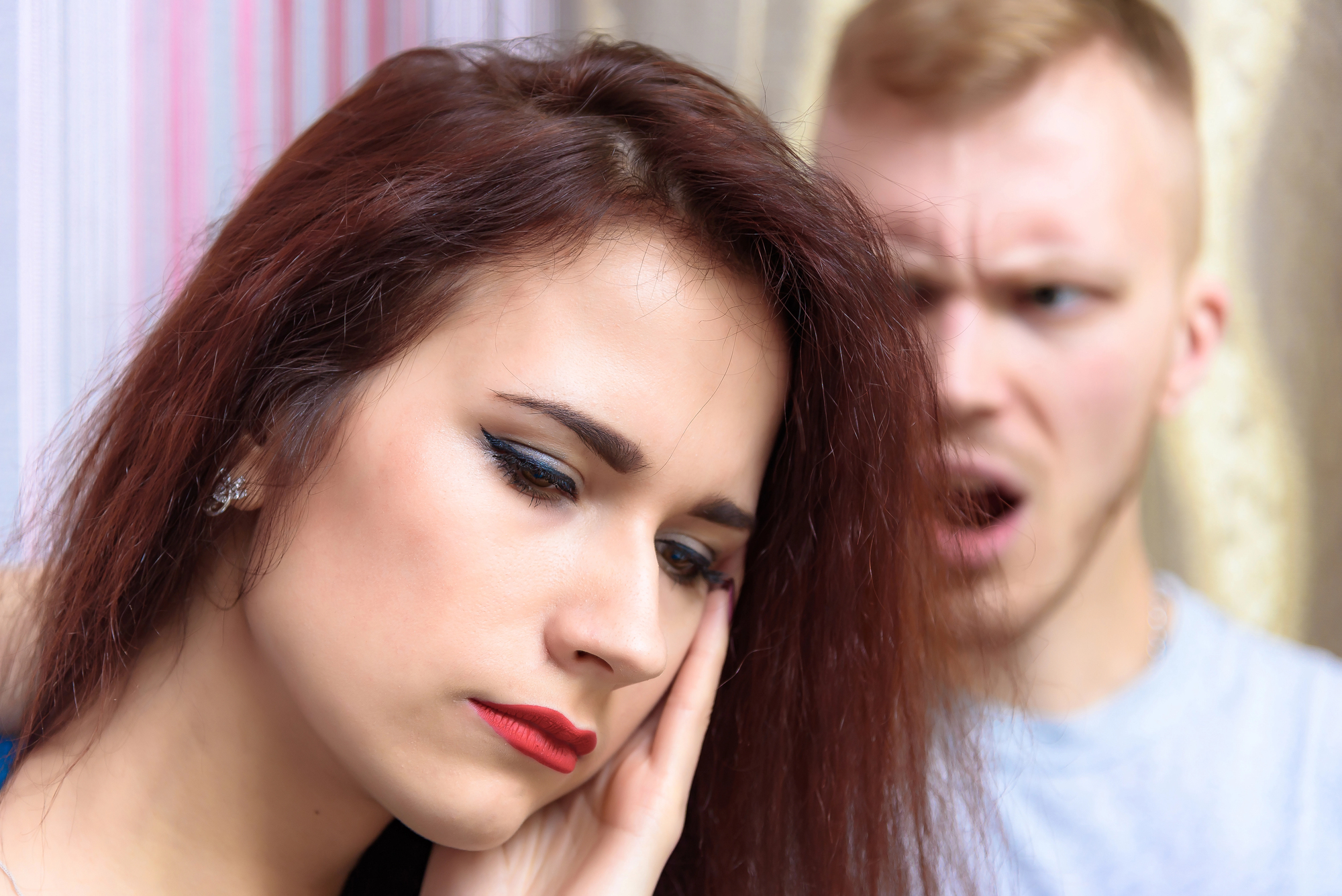 A woman with closed eyes and her hand on her face appears upset. A man in the background looks angry, with his mouth open as if shouting. The scene suggests a tense or emotional moment between the two individuals.