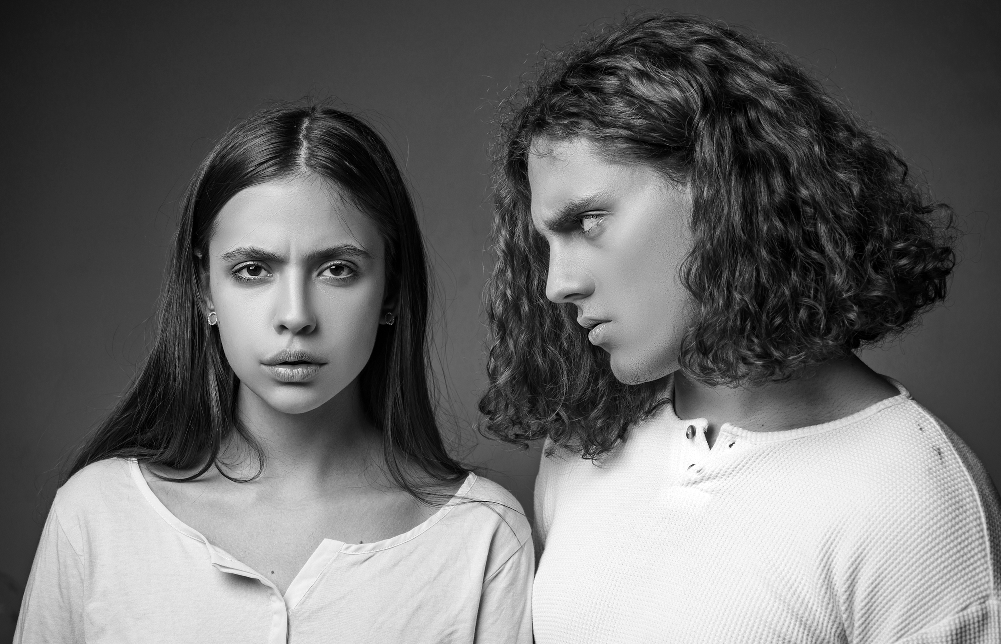 A black and white portrait of a man with curly hair and intense expression looking sideways at a woman with long hair who appears serious, both wearing light-colored tops.