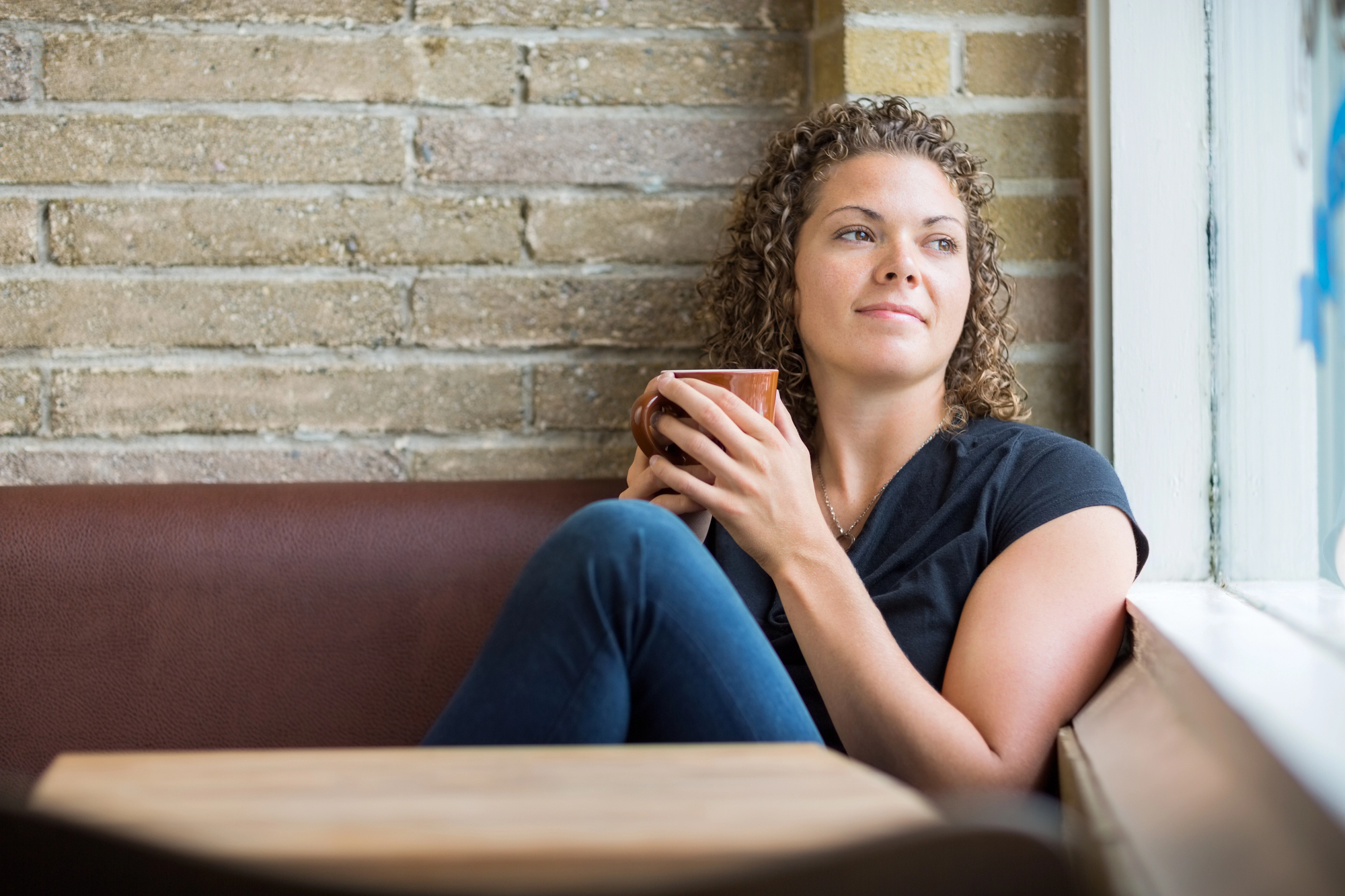 A woman with curly hair sits in a cozy cafe, holding a cup and gazing out the window. She wears a black T-shirt and jeans, relaxed against a brown cushioned bench. The brick wall and wooden table add to the warm ambiance.