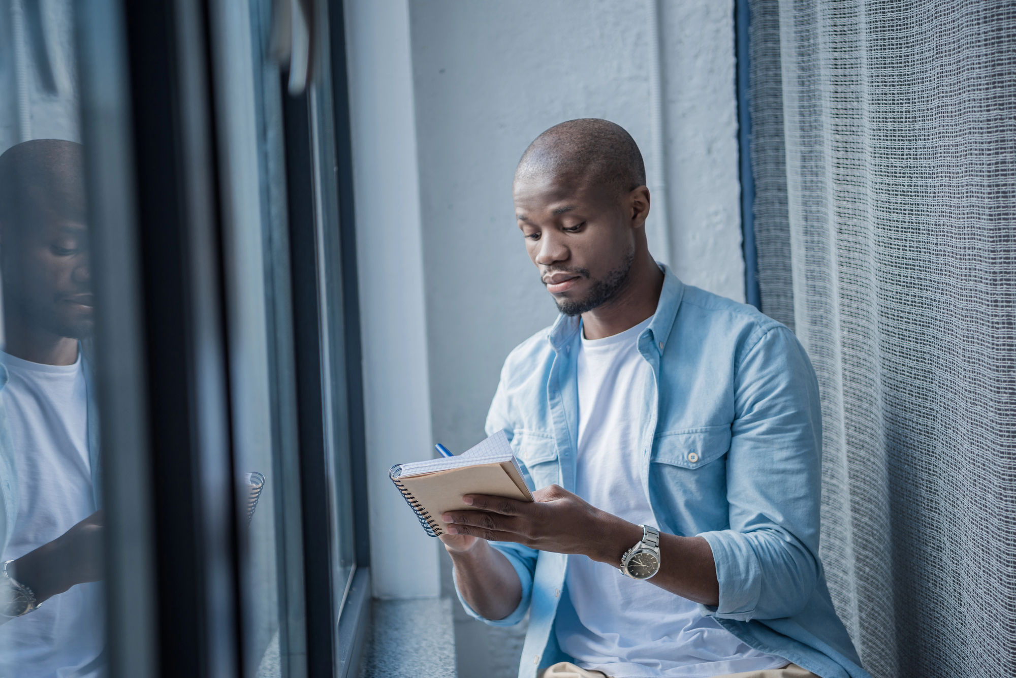 A man in a light blue shirt sits by a window, writing in a notebook. He appears focused and is wearing a watch, with a calm background of curtains and glass reflecting the scene.