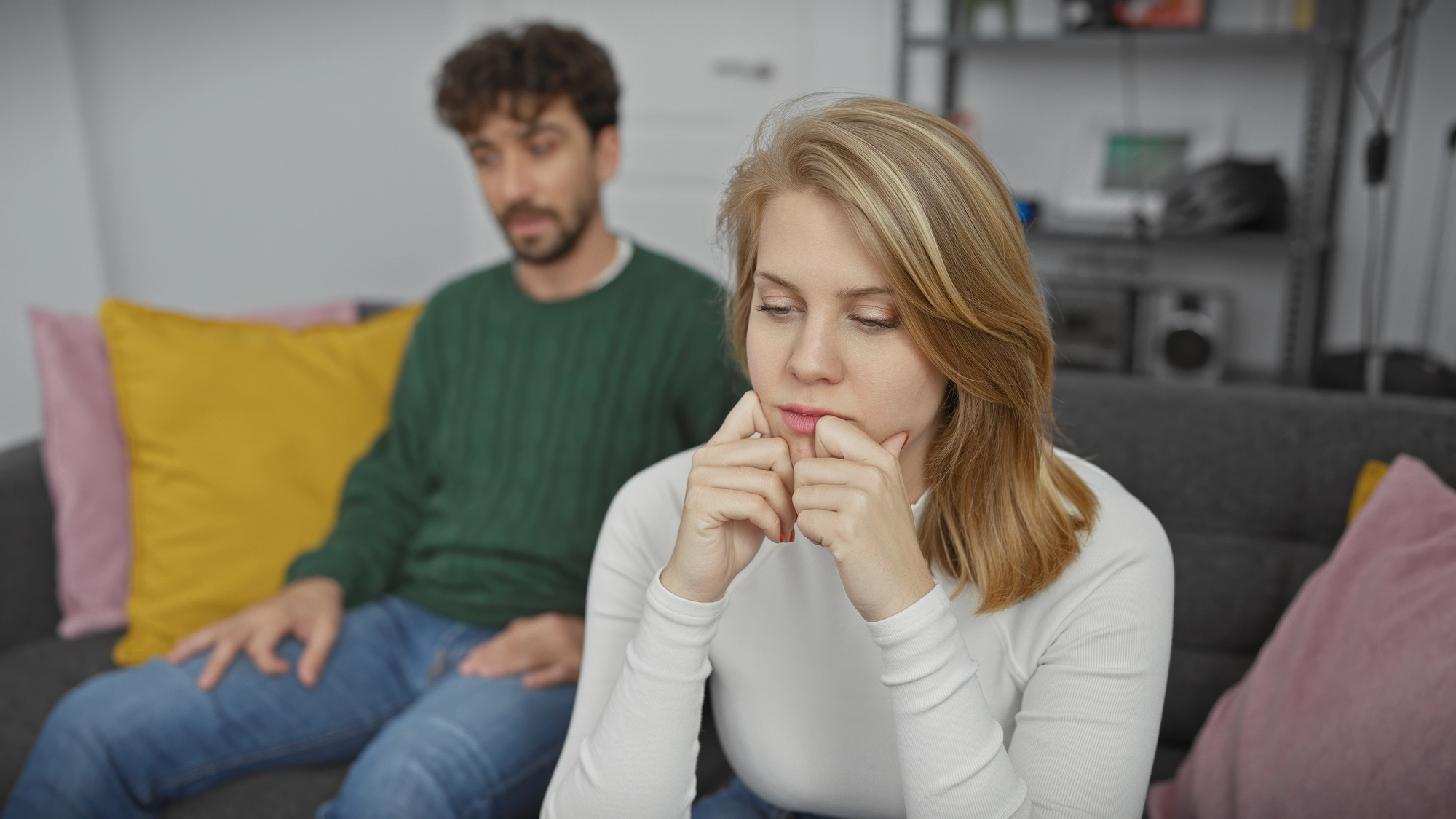 A woman with blonde hair sits on a gray couch, looking pensive and resting her chin on her hands. A man with dark curly hair and wearing a green sweater sits slightly behind her, gazing at her with a concerned expression.