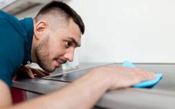 A man with short hair is intently cleaning a stovetop with a blue cloth. The focus is on his concentrated expression as he leans closely to ensure the surface is spotless.