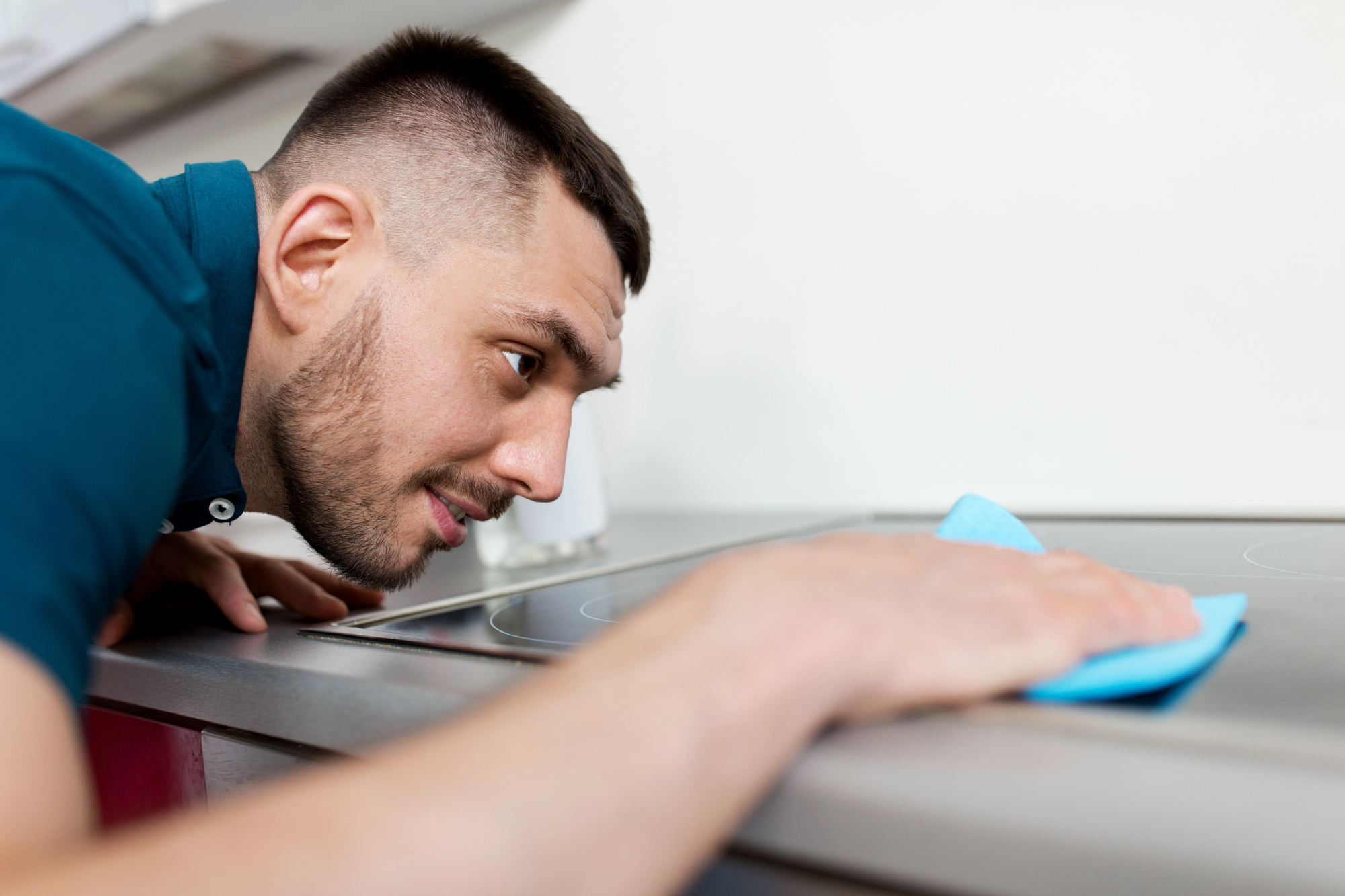 A man with short hair is intently cleaning a stovetop with a blue cloth. The focus is on his concentrated expression as he leans closely to ensure the surface is spotless.