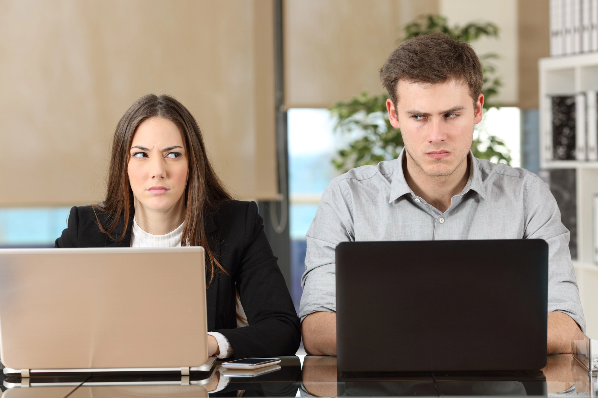 A woman and a man sit side by side at a table, each using a laptop. The woman looks at the man with suspicion, while the man stares intently at his screen. Both appear focused in an office setting.