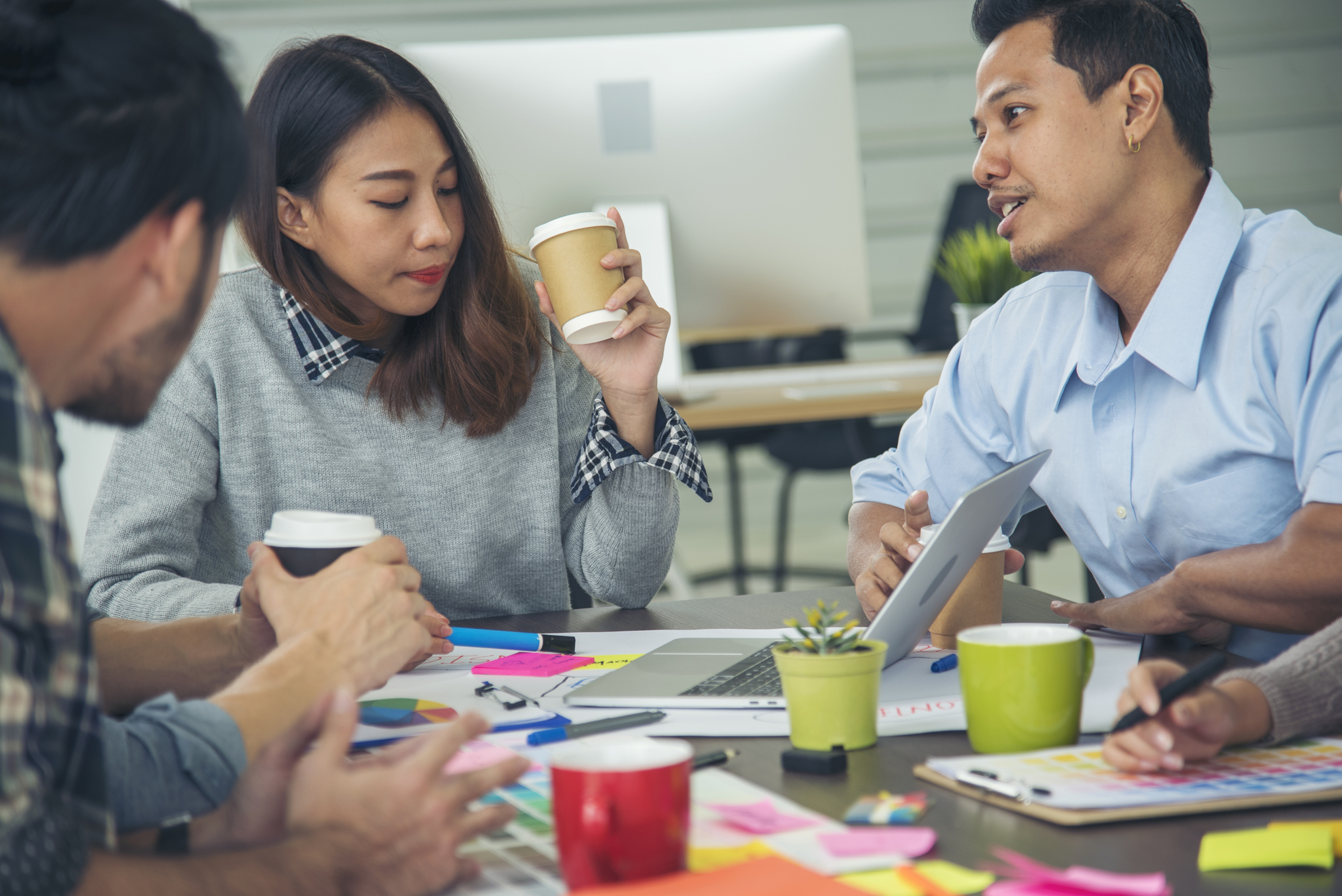 Four people are sitting around a table in an office setting, engaged in a discussion. They have coffee cups, documents, and colorful sticky notes on the table. One person is holding a tablet, and they appear focused and collaborative.