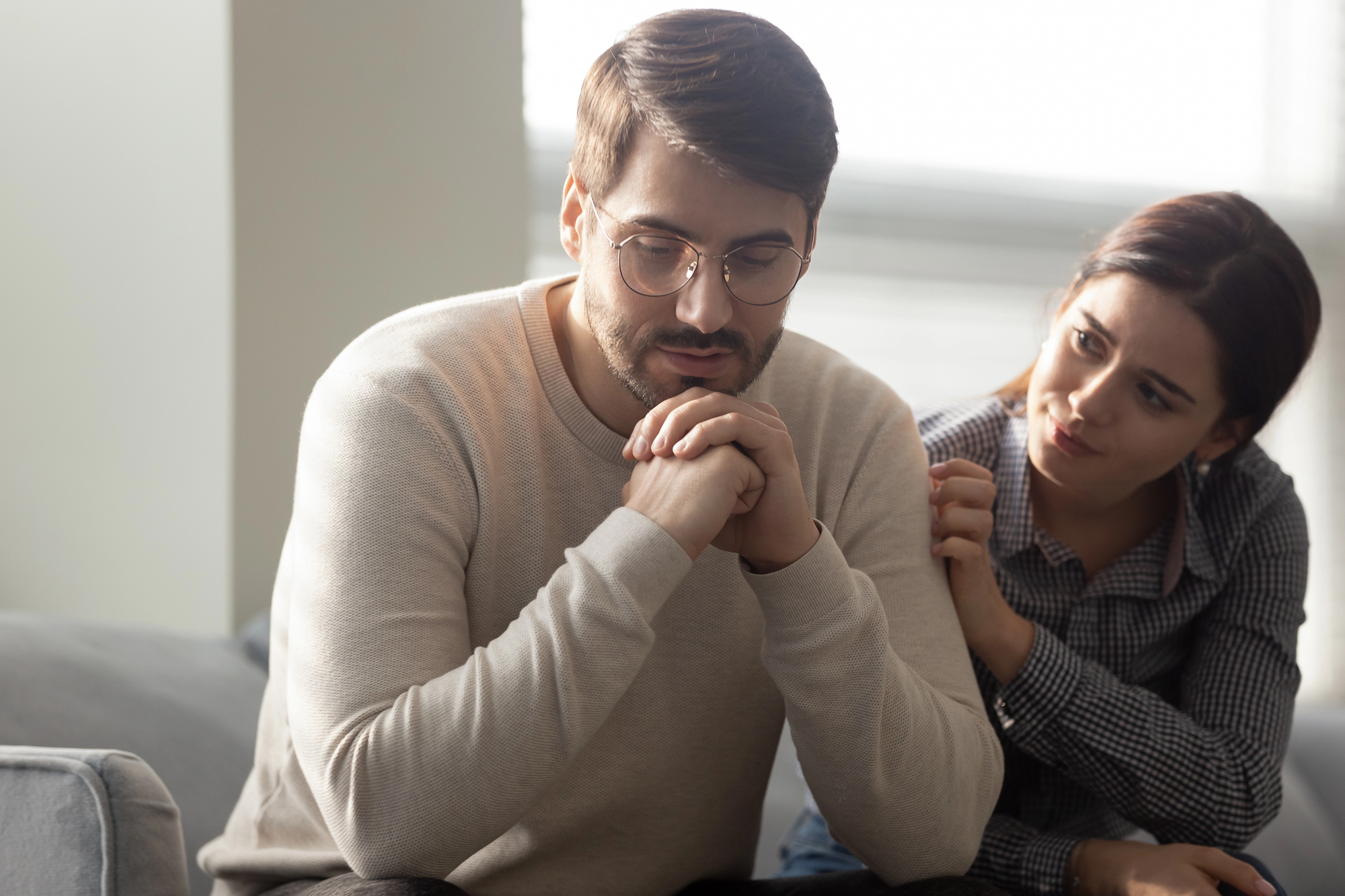 A man with glasses sits on a couch, looking down thoughtfully, with his hands clasped. A woman beside him gently touches his shoulder, offering comfort and support. The room is softly lit with sunlight coming through a window.