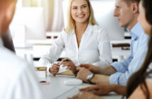 A group of colleagues is engaged in a meeting around a table in a modern office. A smiling woman in a white shirt sits at the center, holding a pen and looking at her coworkers, who are attentively listening. Various office supplies are on the table.