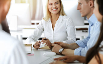 A group of colleagues is engaged in a meeting around a table in a modern office. A smiling woman in a white shirt sits at the center, holding a pen and looking at her coworkers, who are attentively listening. Various office supplies are on the table.