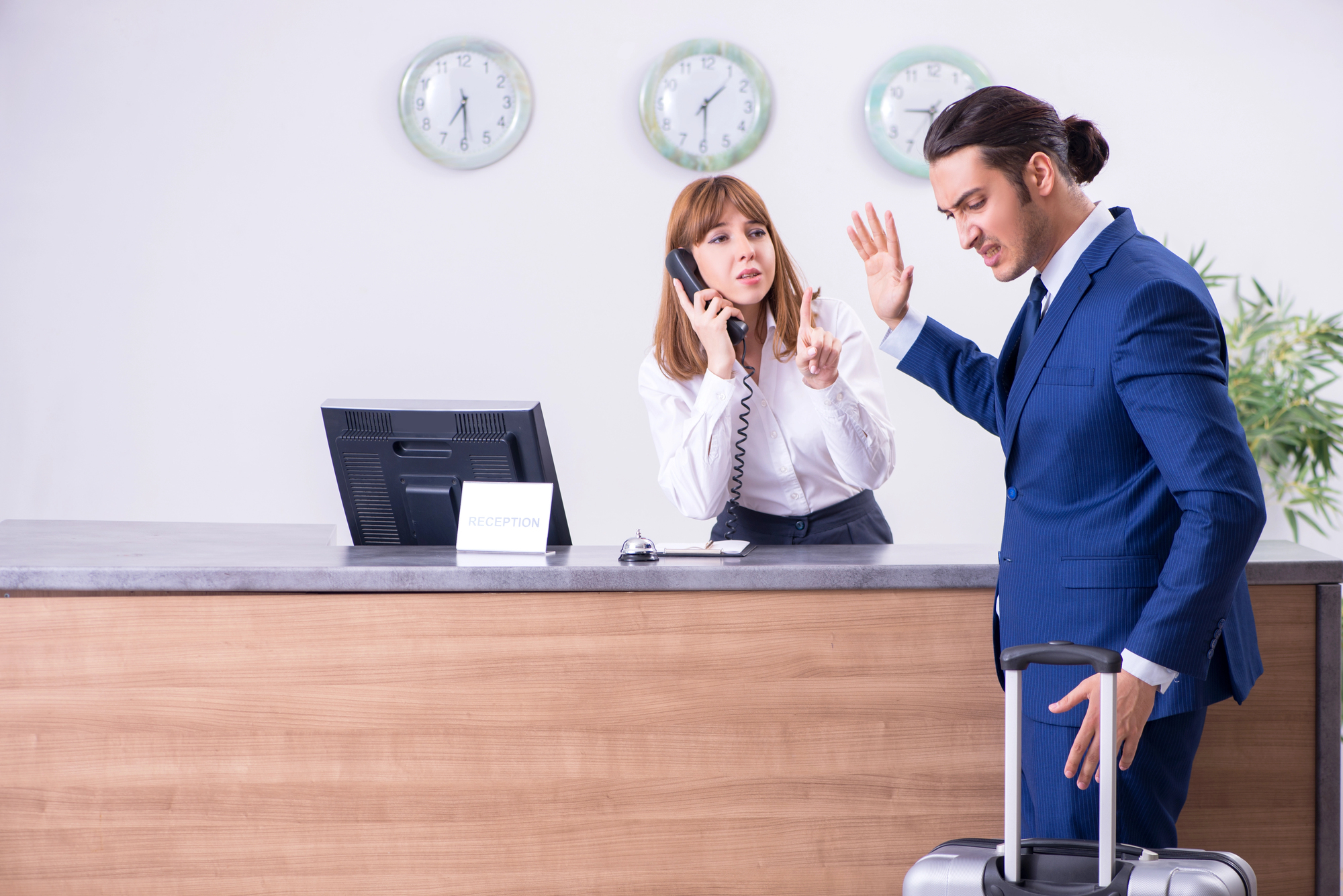 A woman at a reception desk is speaking on the phone and raising her hand towards a man in a blue suit who looks upset and is gesturing with his hand, holding a suitcase. Two clocks are visible on the wall behind them.