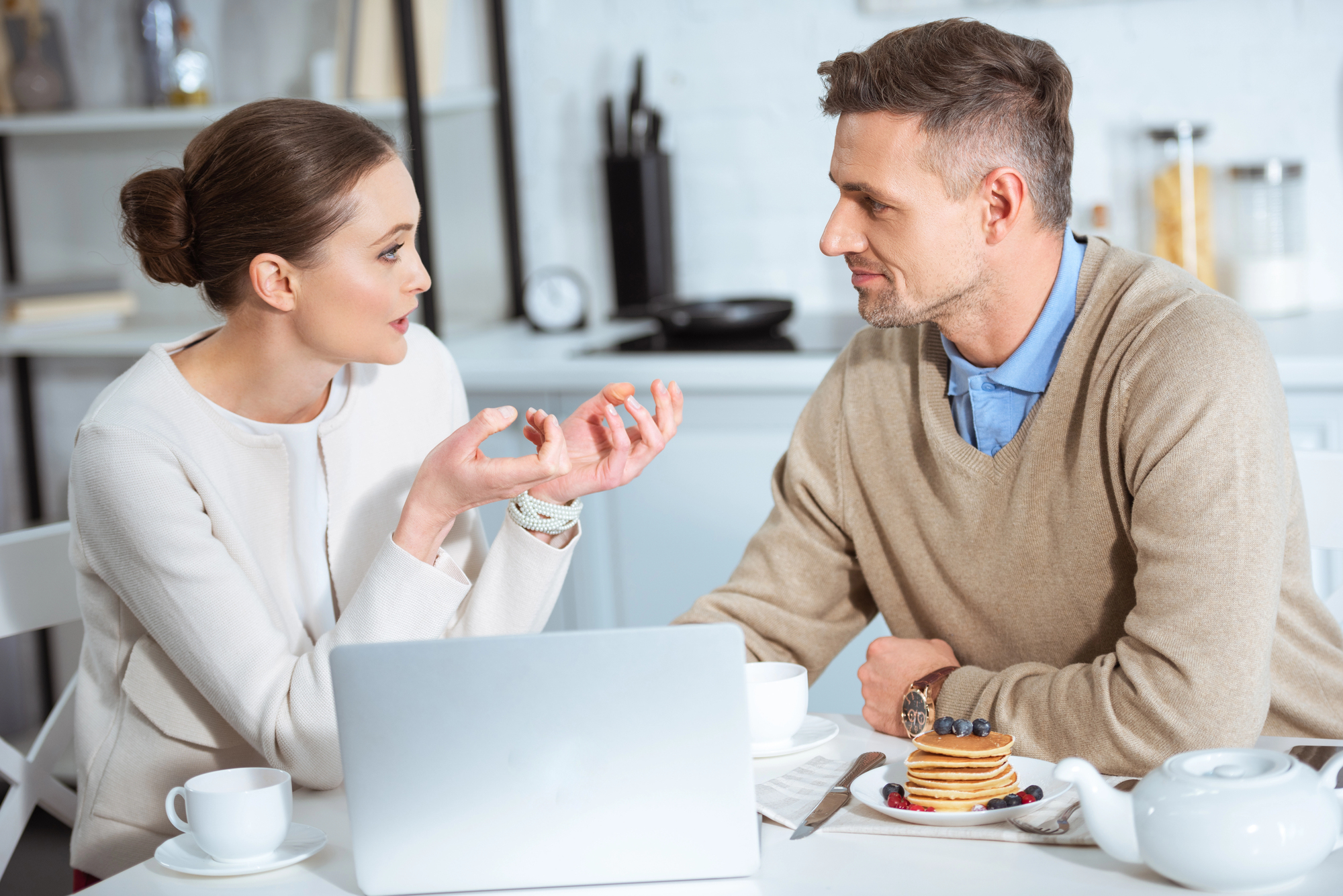 A woman and a man sit at a table with teacups, a laptop, and a stack of pancakes topped with berries. They are engaged in conversation in a bright, modern kitchen. The woman gestures with her hands while the man listens attentively.