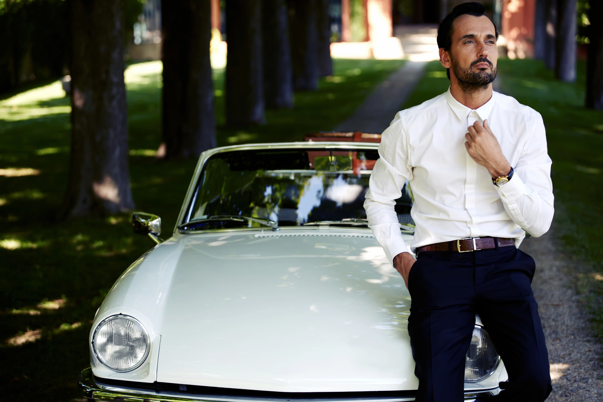 A man in a white shirt and dark pants stands confidently in front of a classic white convertible car on a tree-lined path, adjusting his collar with one hand while the other rests in his pocket.