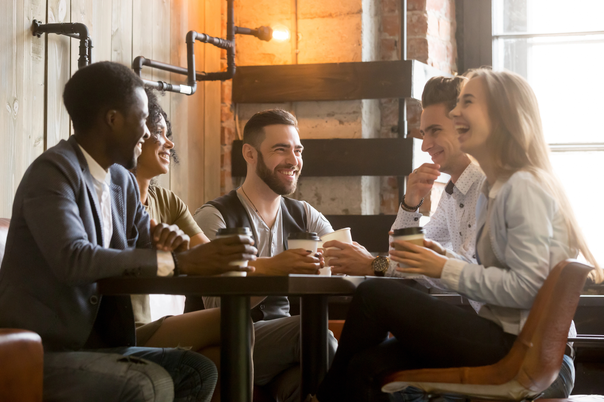 A group of five people, diverse in appearance, are sitting around a table in a cozy, industrial-style coffee shop with brick walls and exposed pipes. They are smiling, laughing, and enjoying their drinks while engaged in conversation.