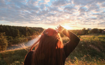 A person with long hair stands in a field at sunset, facing the sun with a hand on their head. Sunlight illuminates their hair, and the sky is filled with scattered clouds. There is a river and dense trees in the background.