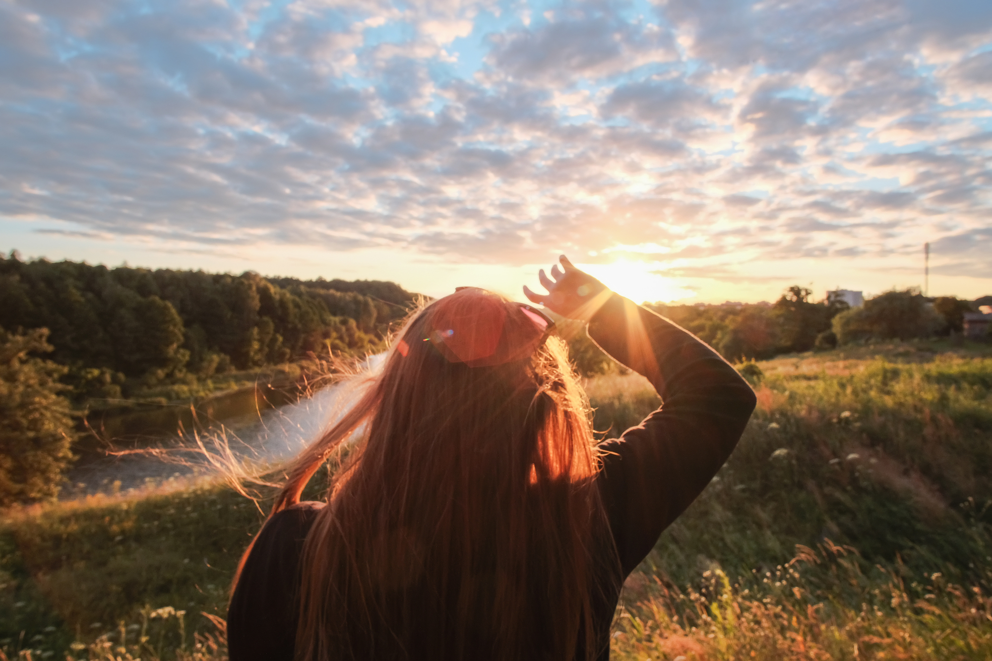 A person with long hair stands in a field at sunset, facing the sun with a hand on their head. Sunlight illuminates their hair, and the sky is filled with scattered clouds. There is a river and dense trees in the background.