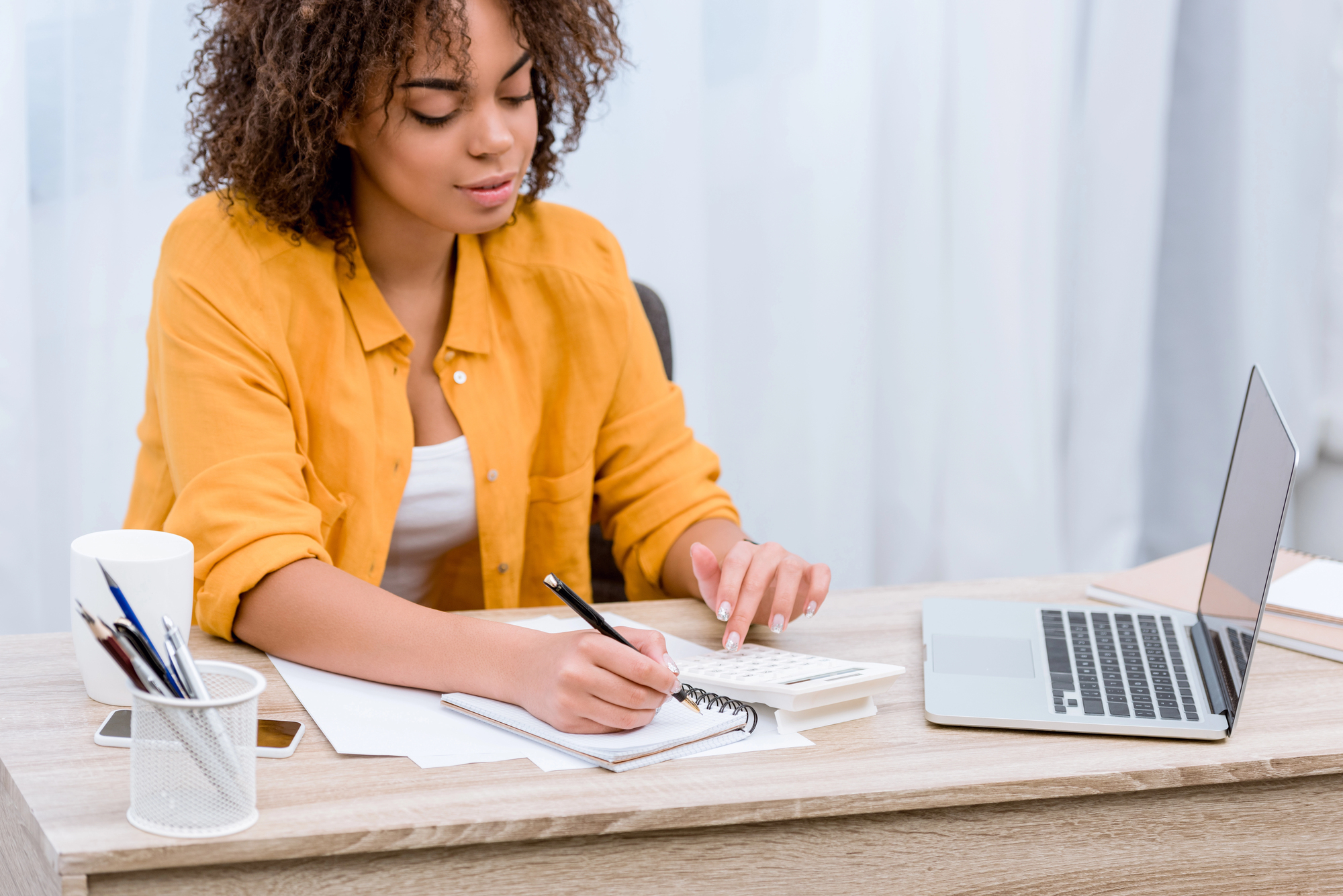 A woman in a yellow blouse sits at a desk, writing in a notebook. She is using a calculator and has a laptop open beside her. The desk also holds a mug with pens. The background features light-colored curtains.