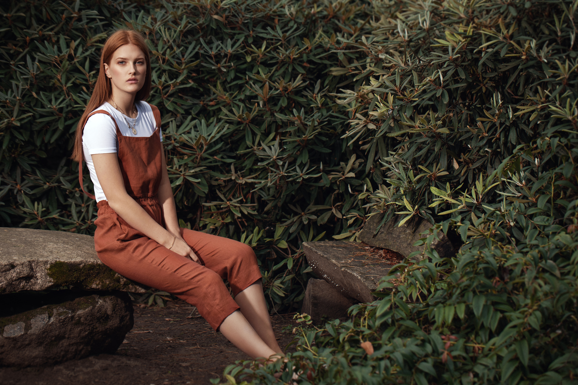 A young woman with long red hair sits on a stone bench in a lush, green garden. She wears a white shirt underneath rust-colored overalls, looking thoughtfully into the distance. Dense bushes and foliage surround her, creating a serene and natural setting.