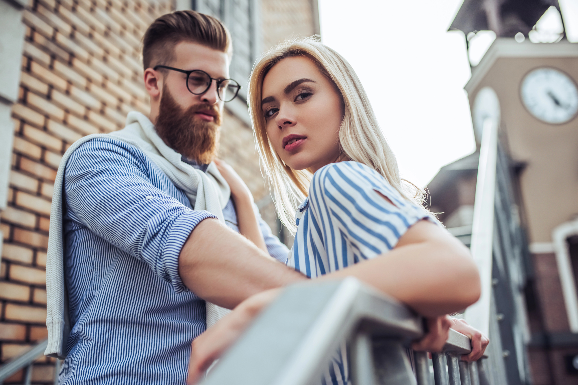 A bearded man with glasses and a blonde woman in a striped blouse pose together on an outdoor staircase with a brick wall and a clock tower in the background. They both have serious expressions and are looking at the camera.