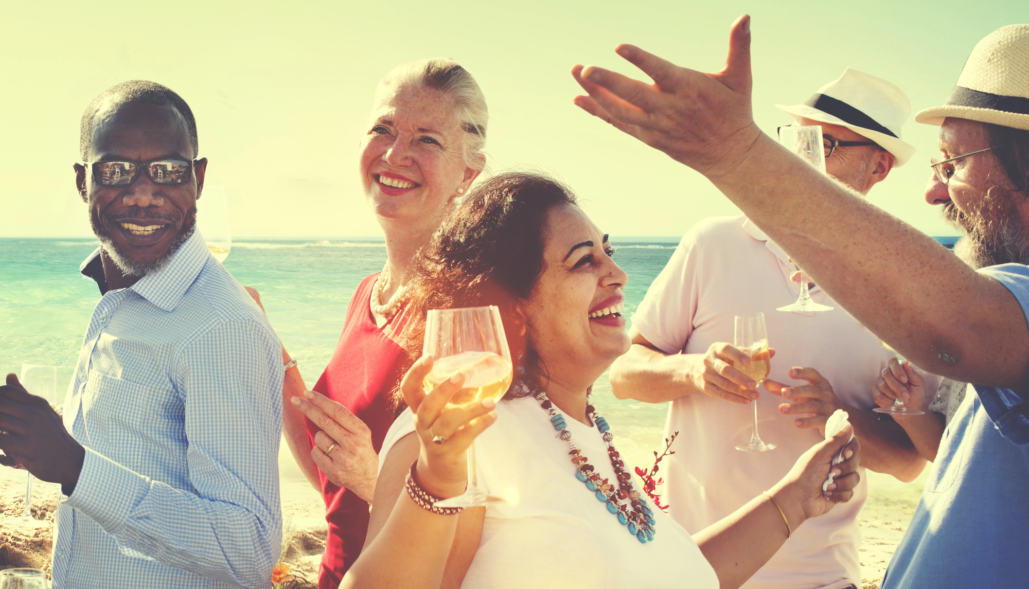 A diverse group of people enjoying a beach party, smiling and holding glasses of wine. The ocean is visible in the background, and everyone appears to be having a good time under the sunny sky.