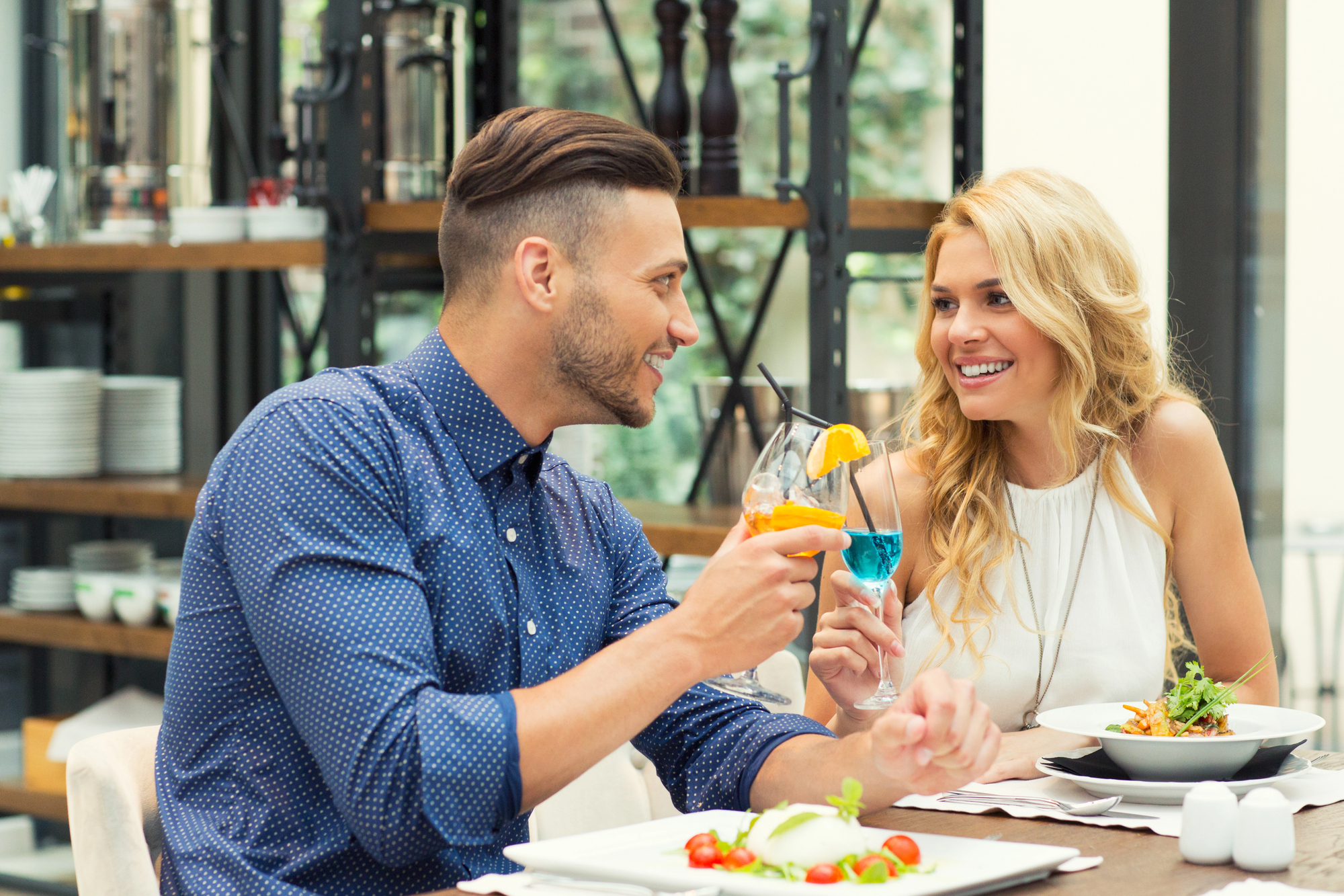 A man and woman are sitting at a restaurant table, smiling and clinking glasses. They are having a meal, with plates of food in front of them. The setting is modern and well-lit, with shelves and tableware in the background.