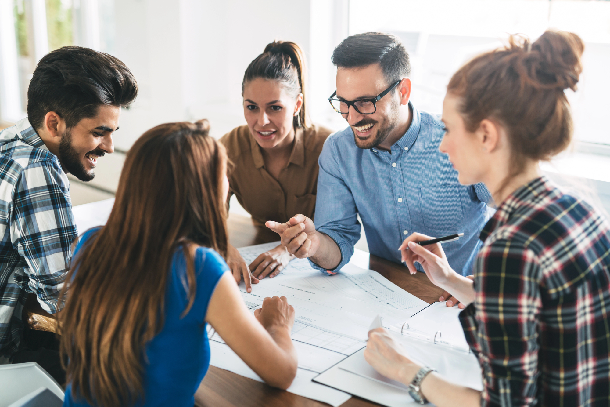 A group of five people sitting around a table, collaborating and discussing. They are looking at documents and blueprints, smiling and engaged in conversation. The atmosphere appears positive and productive.