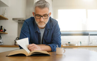 A man with gray hair and glasses wearing a denim shirt is reading a book in a kitchen. He is leaning on a wooden table with a cup beside him. Sunlight is streaming through a window in the background.