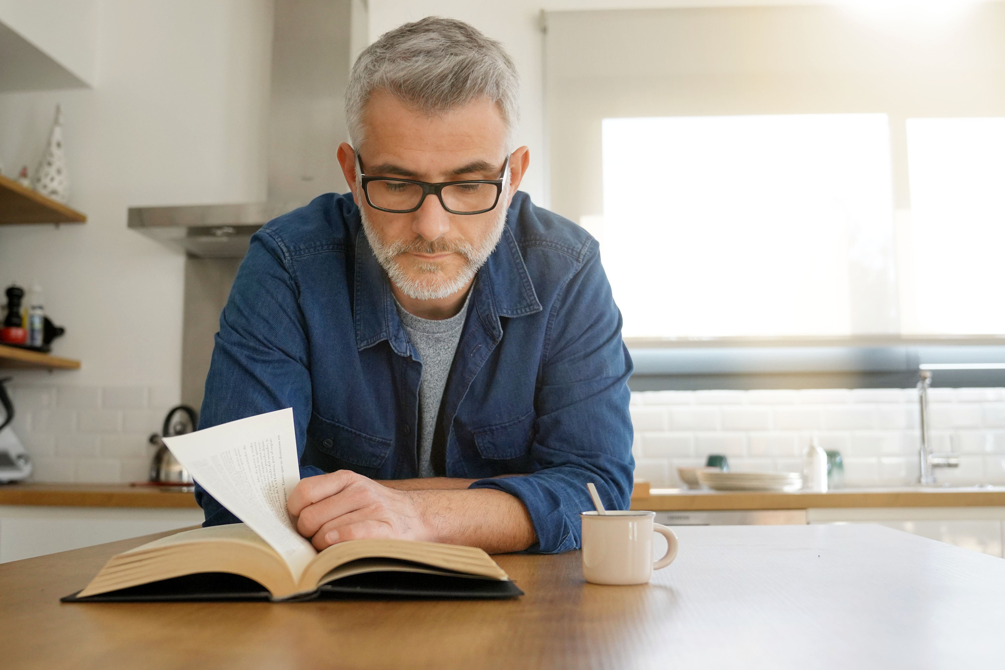 A man with gray hair and glasses wearing a denim shirt is reading a book in a kitchen. He is leaning on a wooden table with a cup beside him. Sunlight is streaming through a window in the background.