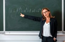 A woman in professional attire stands in front of a chalkboard, pointing towards a mathematical equation involving (a + b)². She wears glasses and appears to be explaining the equation during a lesson. The classroom setting suggests she might be a teacher or professor.