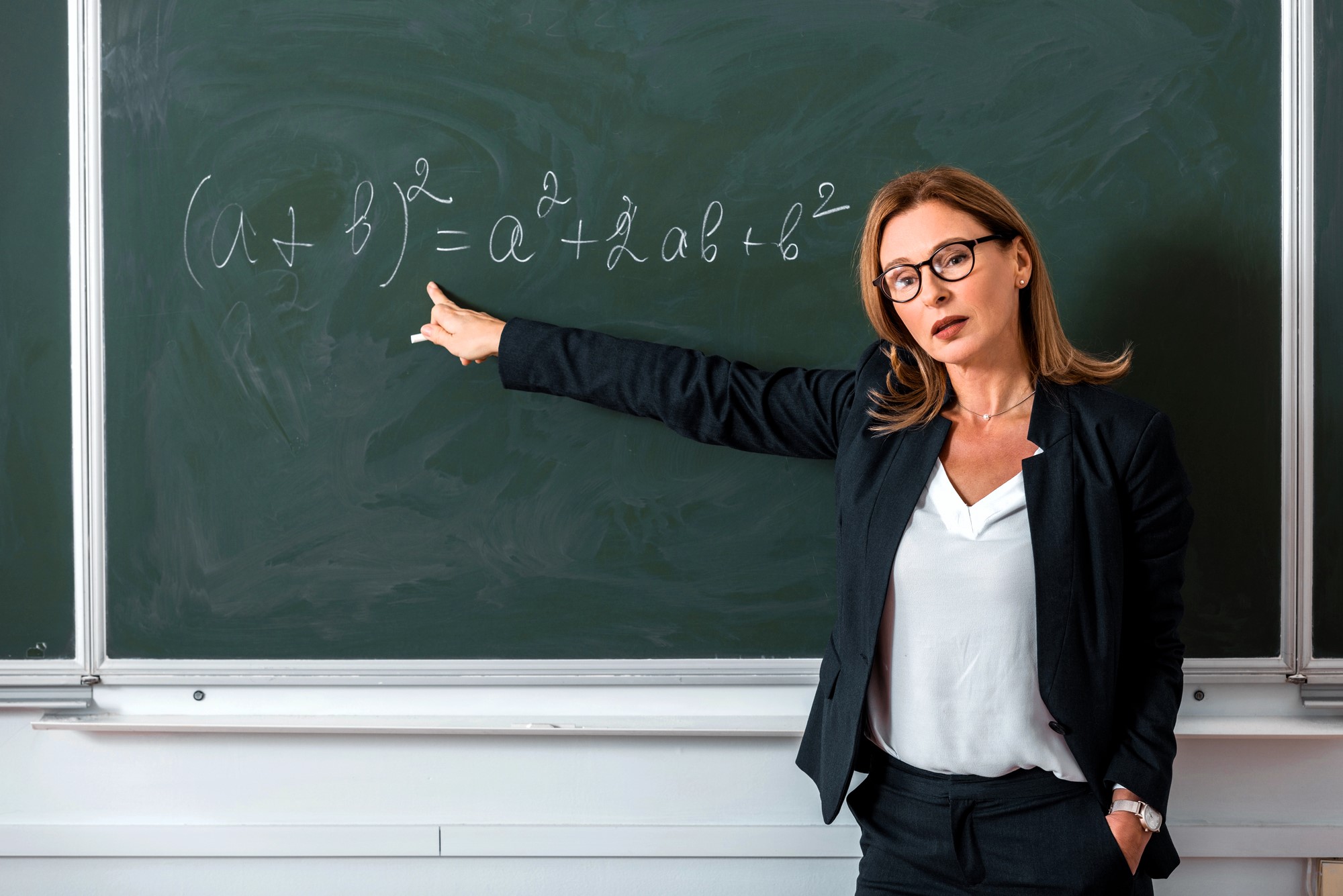 A woman in professional attire stands in front of a chalkboard, pointing towards a mathematical equation involving (a + b)². She wears glasses and appears to be explaining the equation during a lesson. The classroom setting suggests she might be a teacher or professor.