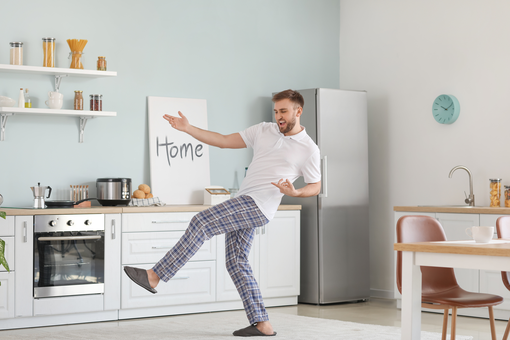 A man in a white shirt and plaid pajama pants dances joyfully in a modern kitchen. The kitchen has white cabinets, a stainless steel fridge, and wall shelves with kitchen items. A sign with the word "Home" is visible on the wall.