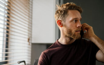 A man with a beard is talking on the phone. He is wearing a dark t-shirt and standing near a window with blinds, allowing soft light to illuminate the room. The background features a white cabinet and dark walls.