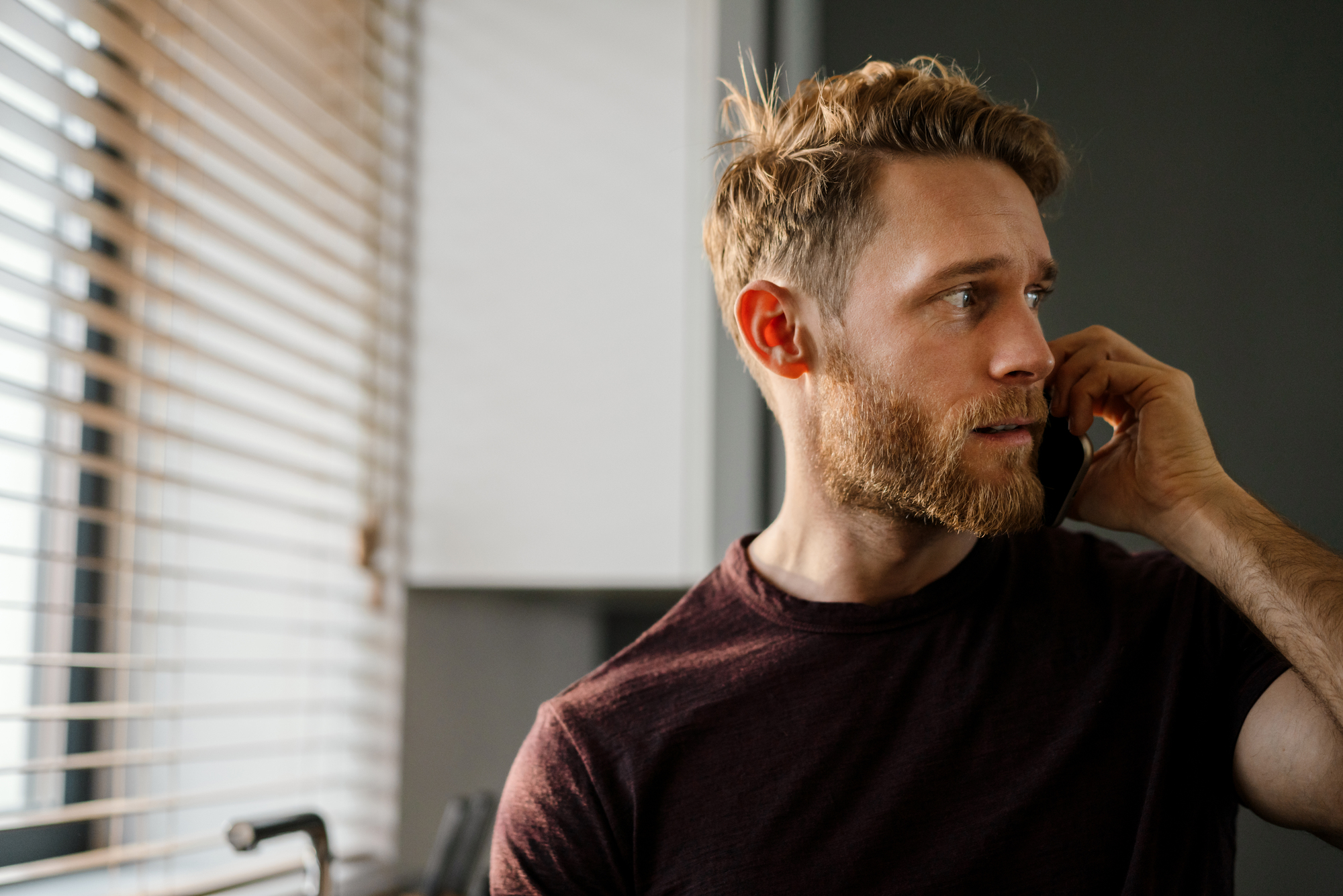 A man with a beard is talking on the phone. He is wearing a dark t-shirt and standing near a window with blinds, allowing soft light to illuminate the room. The background features a white cabinet and dark walls.