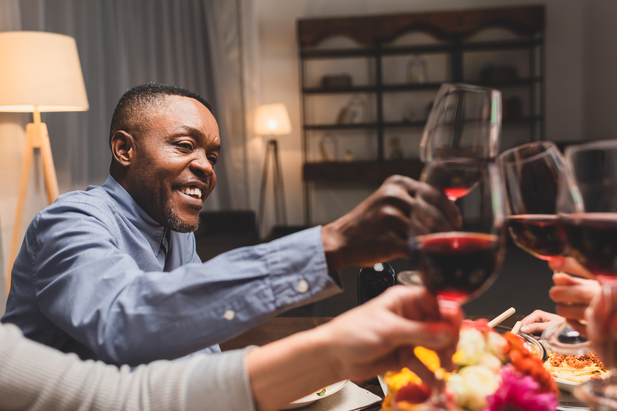 A man in a blue shirt is smiling and clinking glasses of red wine with others at a dining table. The background is warmly lit with lamps and shelves. A bouquet of flowers is visible on the table.