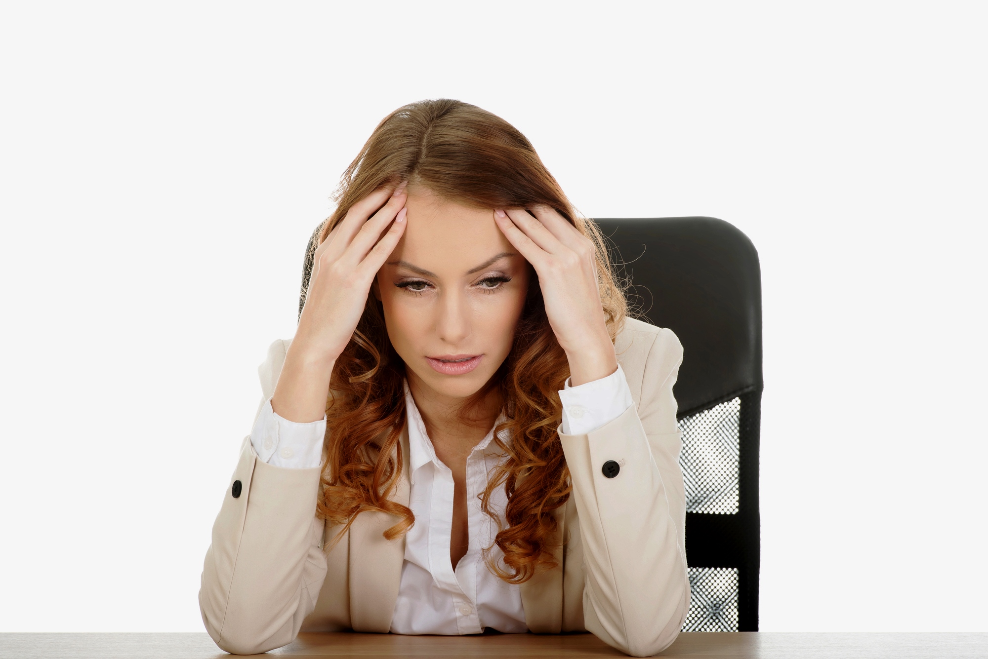 A woman with long, curly hair is seated at a desk, looking stressed. She's wearing a beige blazer and resting her elbows on the desk, with her hands on her temples.