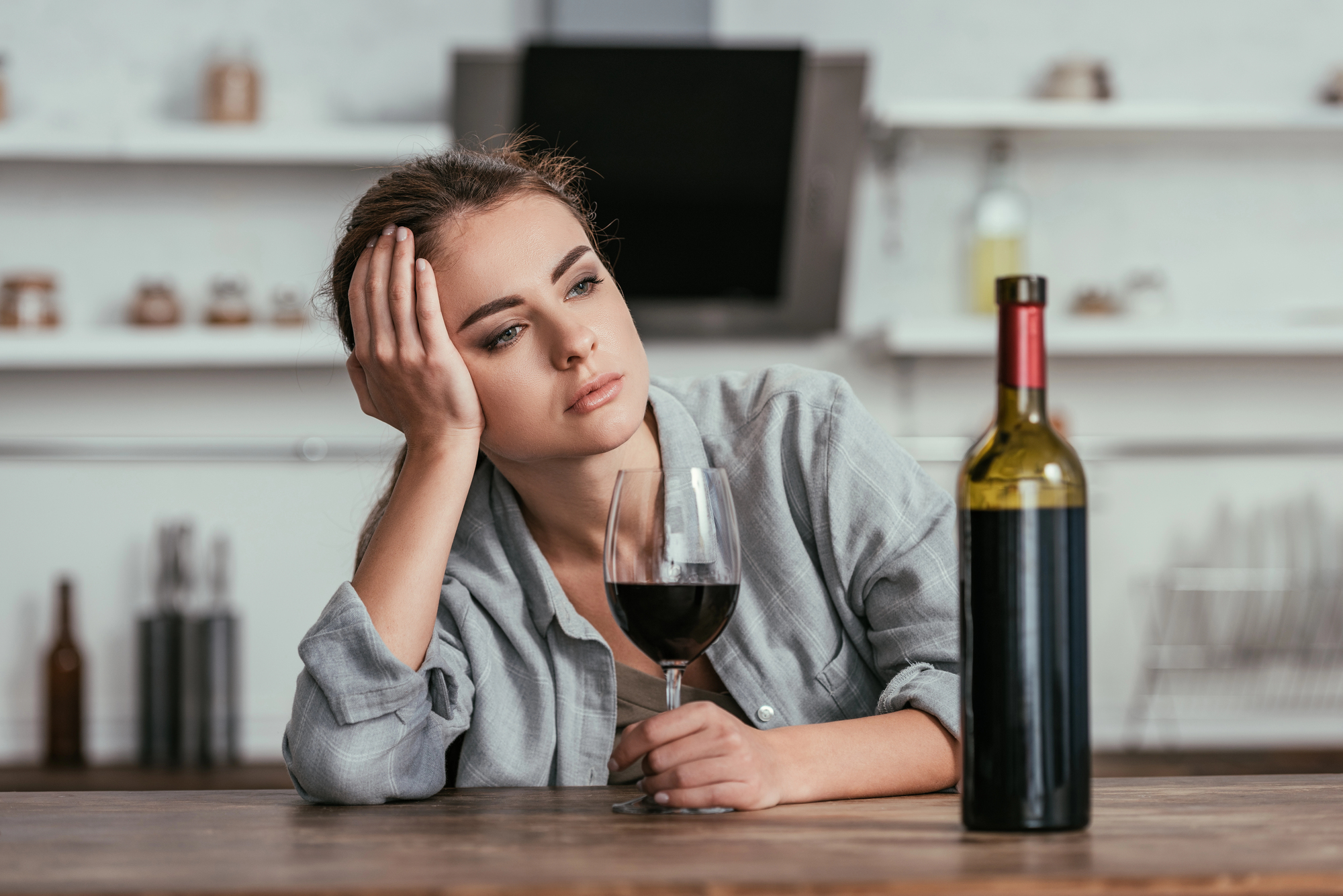 A woman sits at a wooden table, resting her head on her hand while holding a glass of red wine. She has a contemplative expression, and an unopened bottle of wine is also on the table. The background shows a kitchen with shelves and various items.