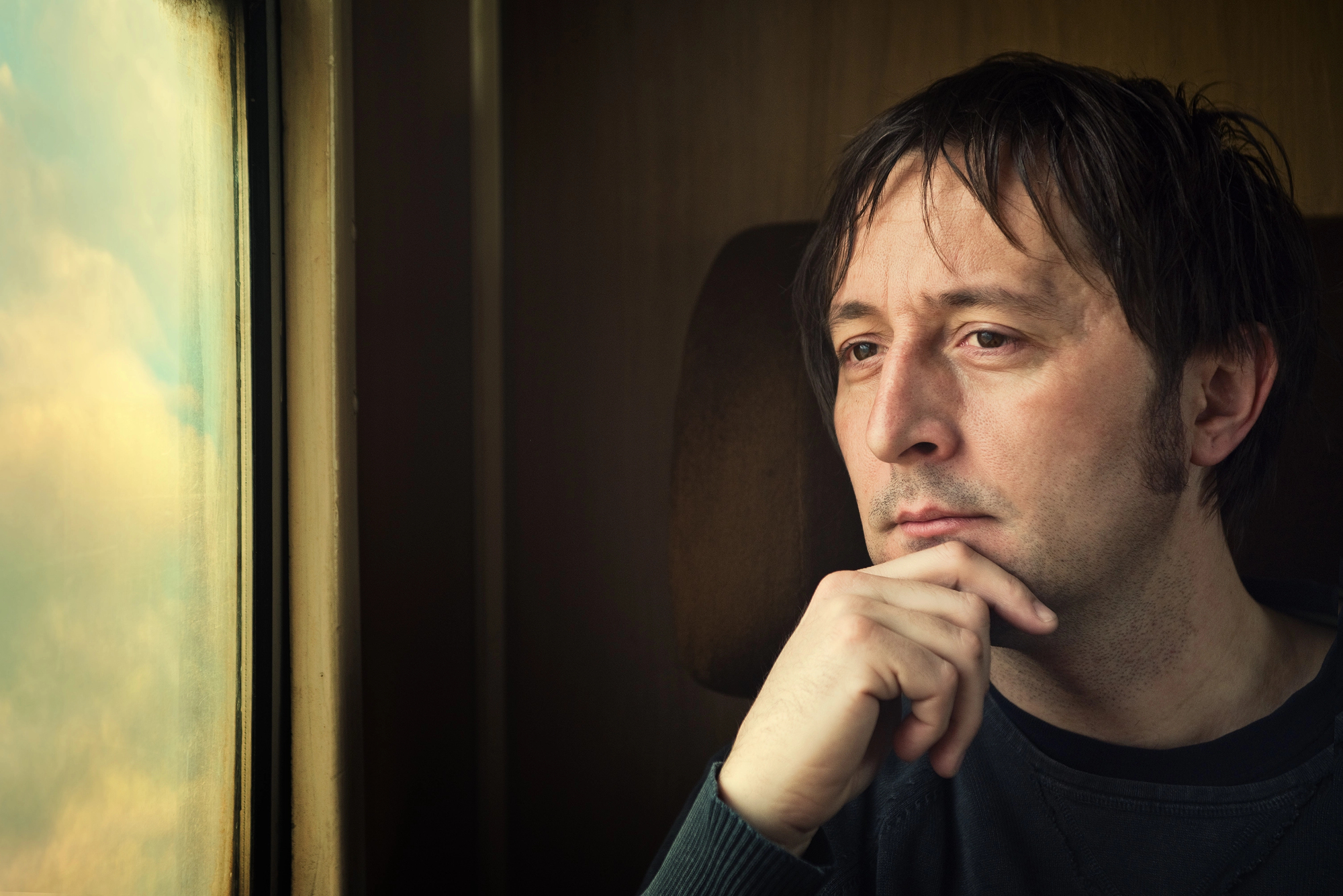 A man gazes thoughtfully out of a train window, resting his chin on his hand. The light from the window casts a soft glow on his face, and the sky outside displays soft, muted colors.