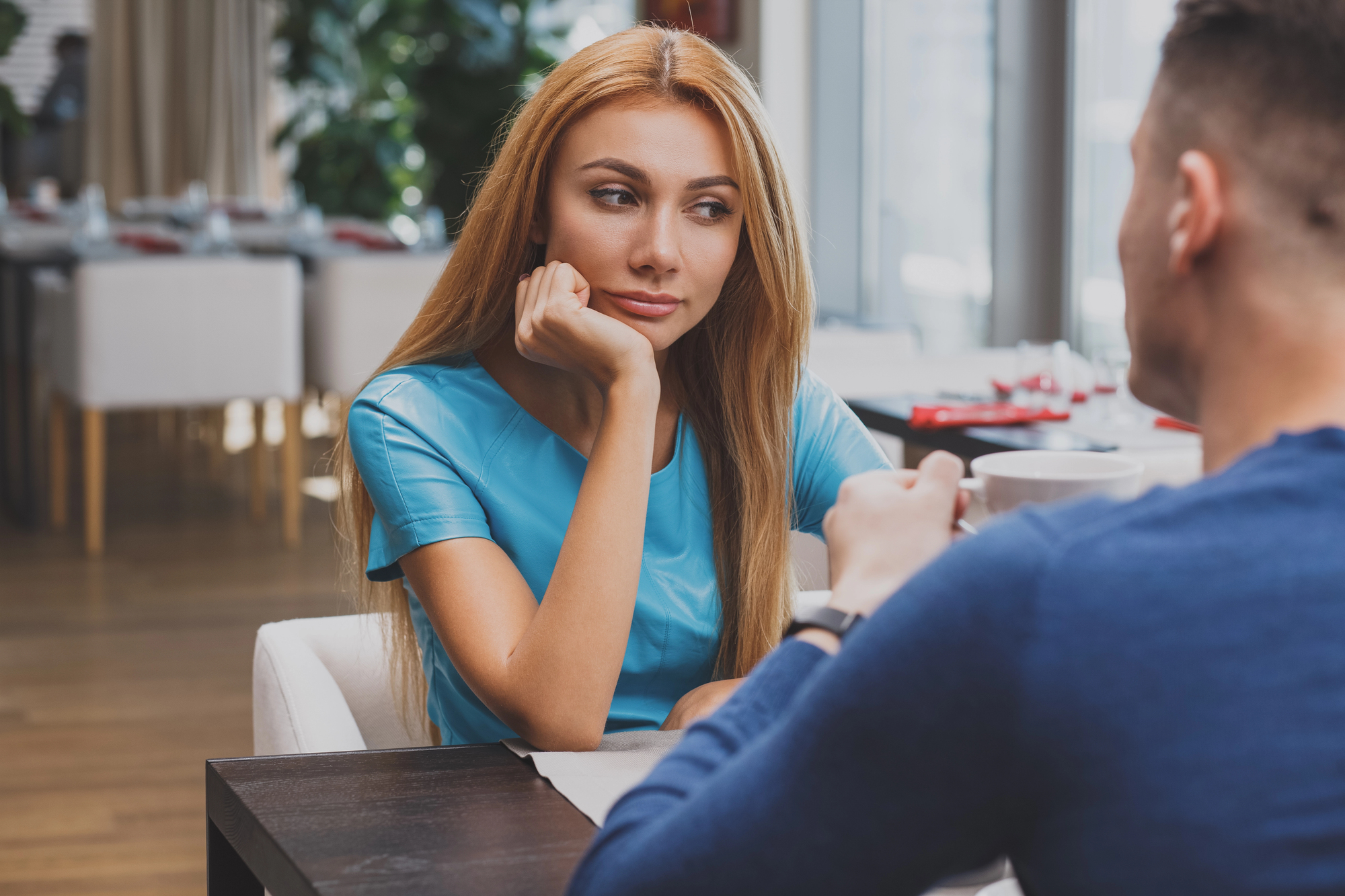 A woman with long blonde hair wearing a blue top rests her chin on her hand, looking at a man across from her at a table in a modern cafe. The man, wearing a blue shirt, faces away from the camera, and they both have drinks in front of them.