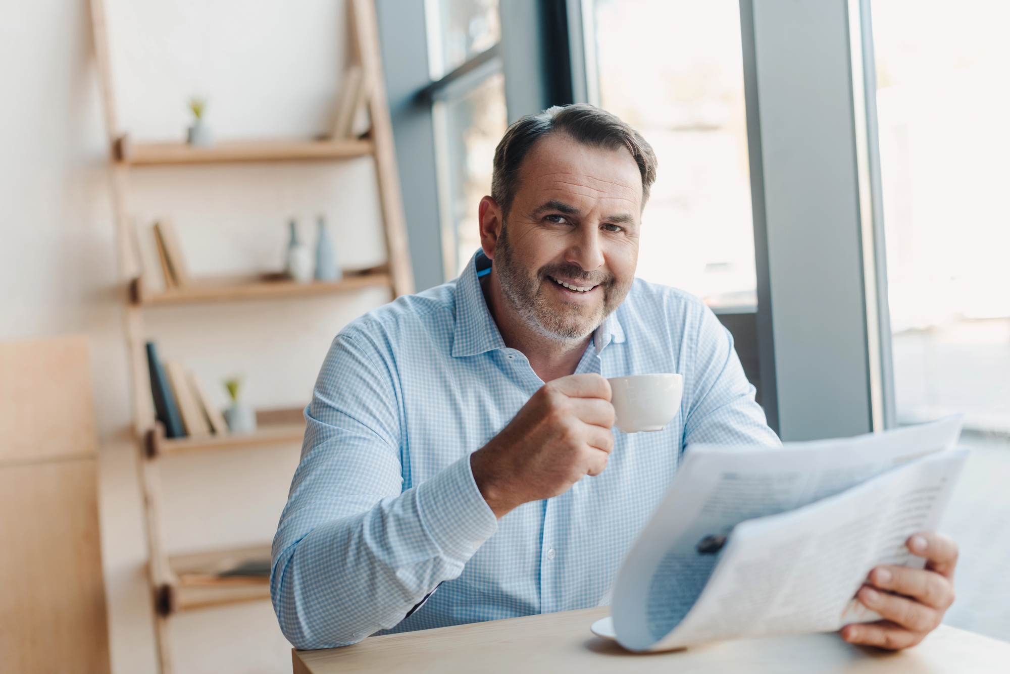 A man in a light blue shirt is sitting at a wooden table, holding a cup of coffee in one hand and a newspaper in the other. He is smiling and sitting in a bright room with a bookshelf in the background.