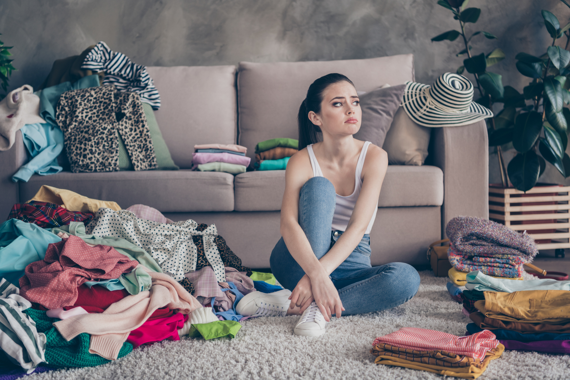 A woman sits on a carpeted floor, surrounded by piles of clothes in a living room. She looks contemplatively into the distance. The couch behind her is also covered with clothes, and there are plants and a striped hat in the background.